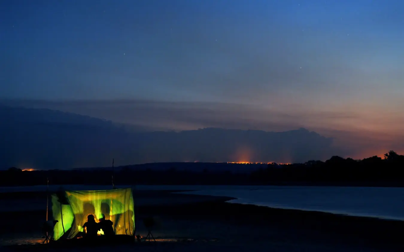 Silhouetted figures in illuminated tent beside lake at dusk, Kiba Point, Nyerere National Park, Tanzania
