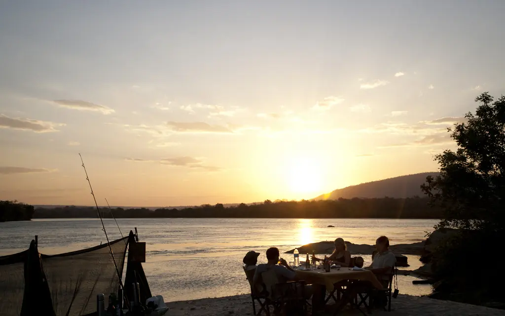 Guests dining at riverside table during golden sunset with fishing boat, Kiba Point, Nyerere National Park, Tanzania