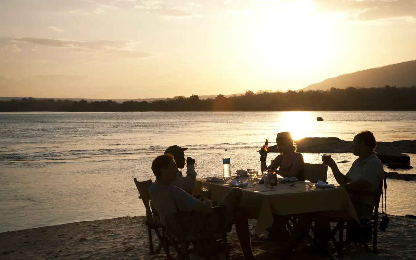 Group dining at table on riverbank at sunset, Kiba Point, Nyerere National Park, Tanzania