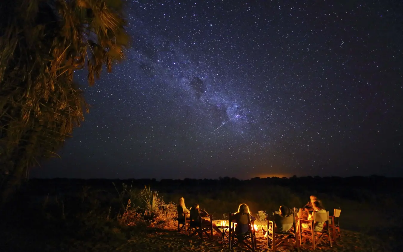 Guests gathered around campfire under starlit sky, Kichaka Expeditions, Ruaha National Park, Tanzania