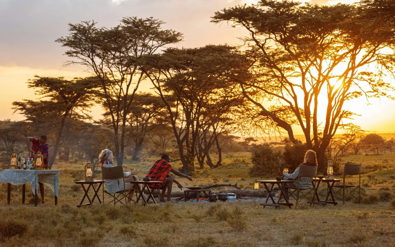 Guests dining outdoors at sunset under acacia trees, Kicheche Valley Naboisho, Maasai Mara, Kenya