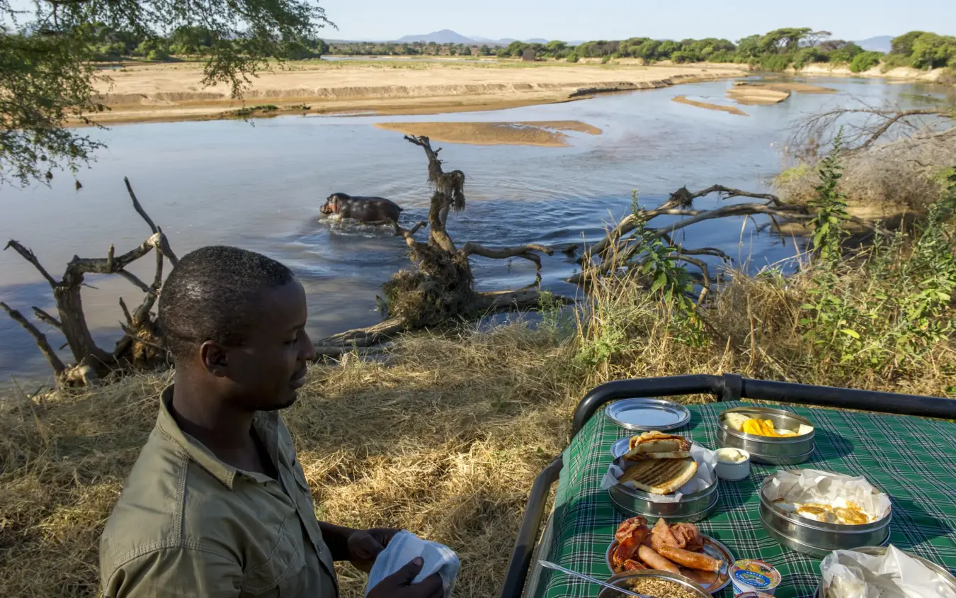 Guide enjoys riverside meal overlooking African savanna landscape, Kigelia Ruaha, Ruaha National Park, Tanzania