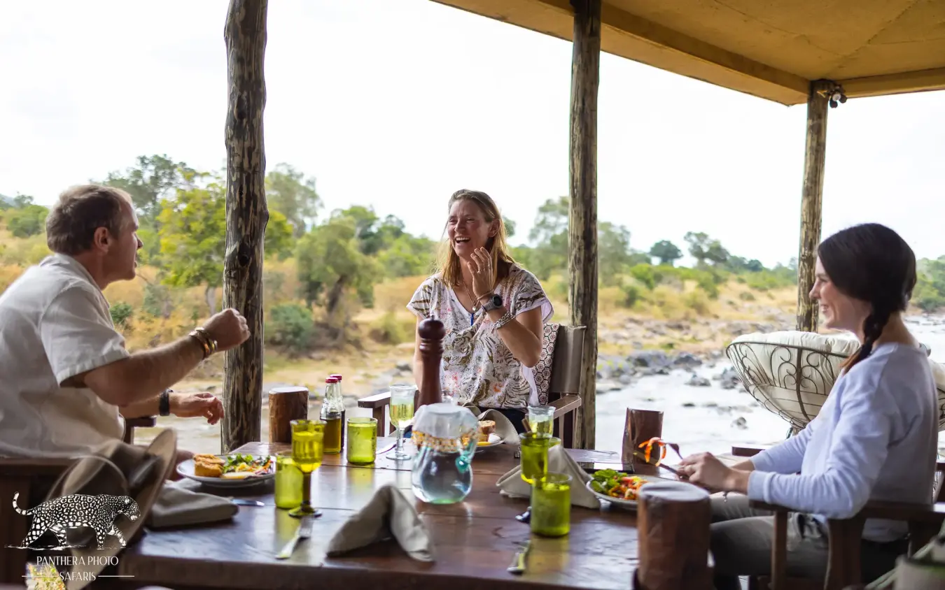 Guests dining at riverside deck overlooking river and bush, Kimya Kimya Camp, Maasai Mara, Kenya