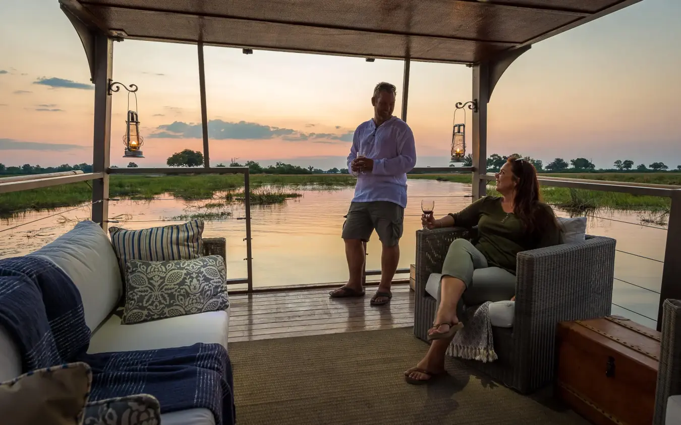 Guests relaxing on covered deck overlooking flooded delta at sunset, King’s Pool, Linyanti Wildlife Reserve, Botswana