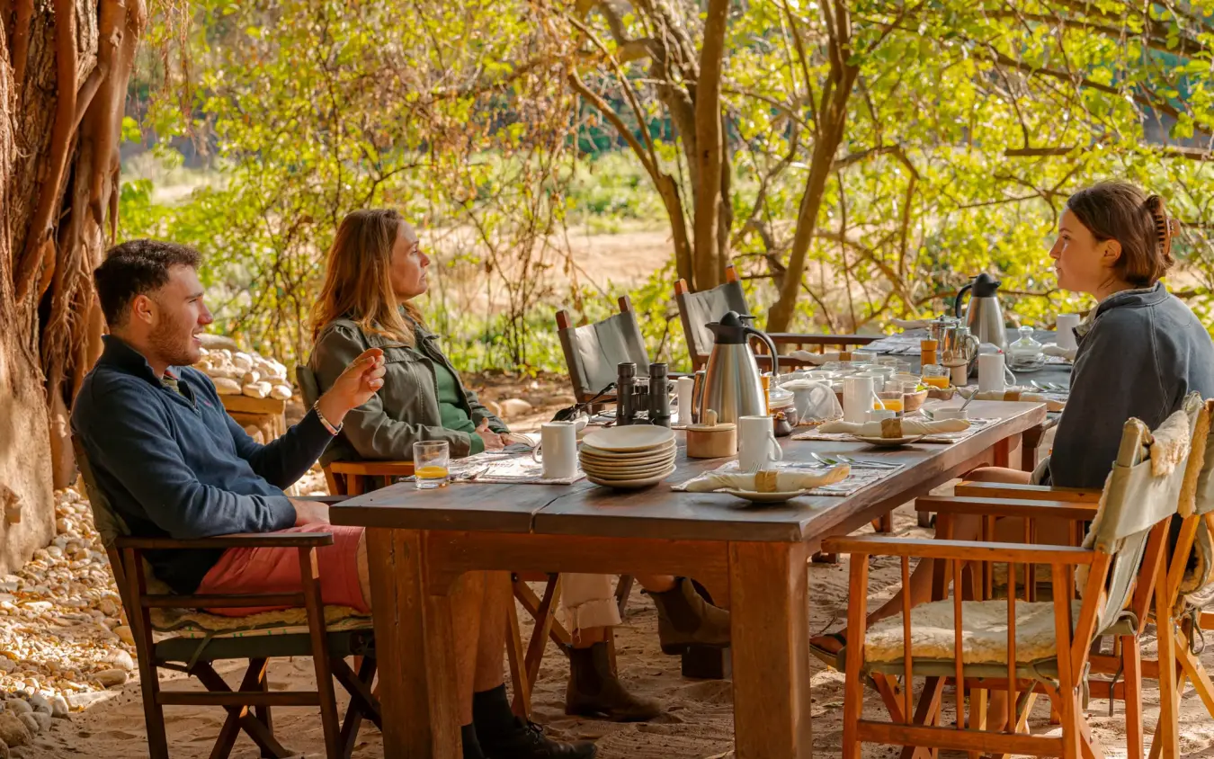 Three guests dining at outdoor table under tree canopy, Kitich Camp, Northern Kenya, Kenya