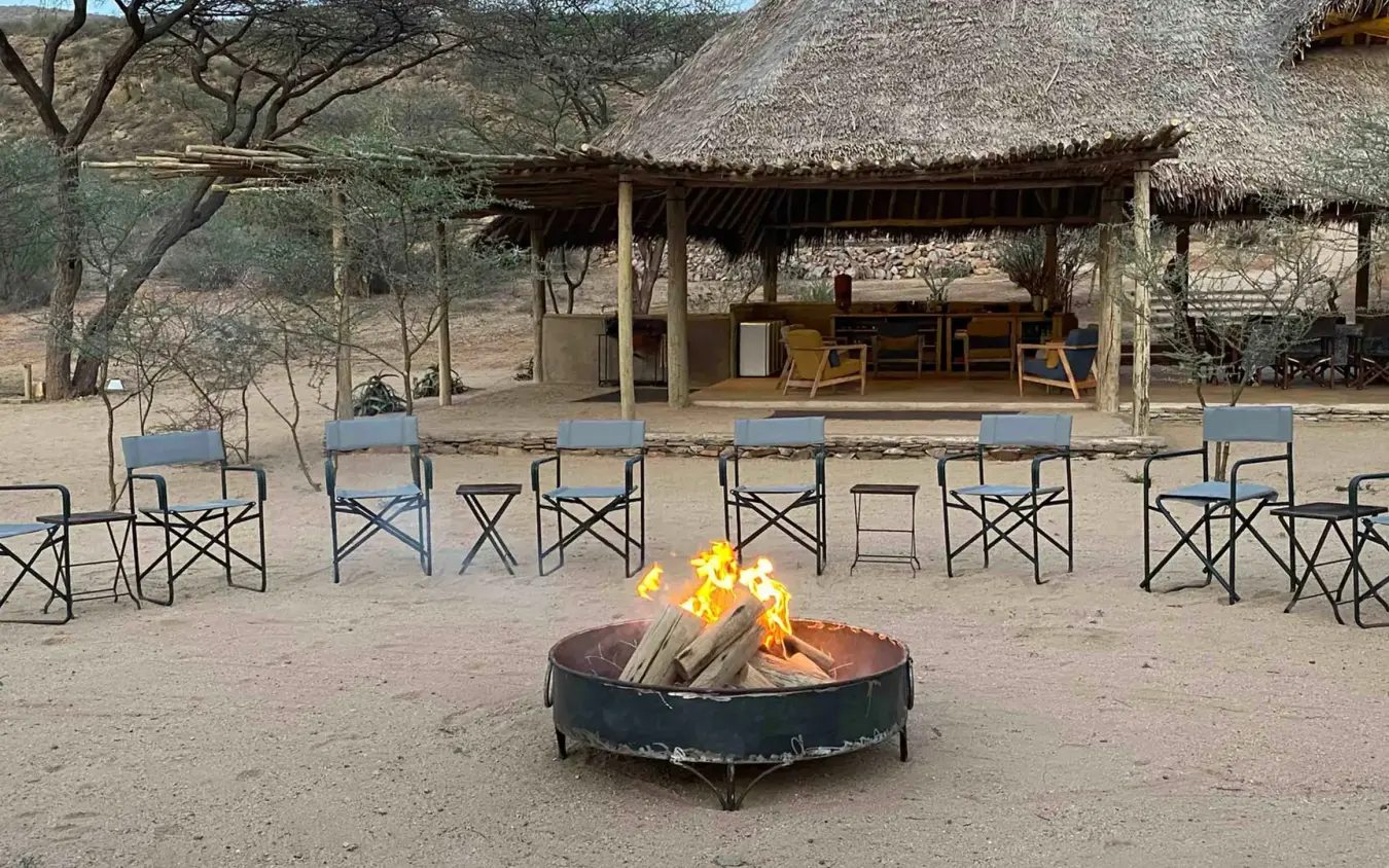 Central firepit with thatched-roof structures and camp chairs, Koros Camp, Northern Kenya, Kenya