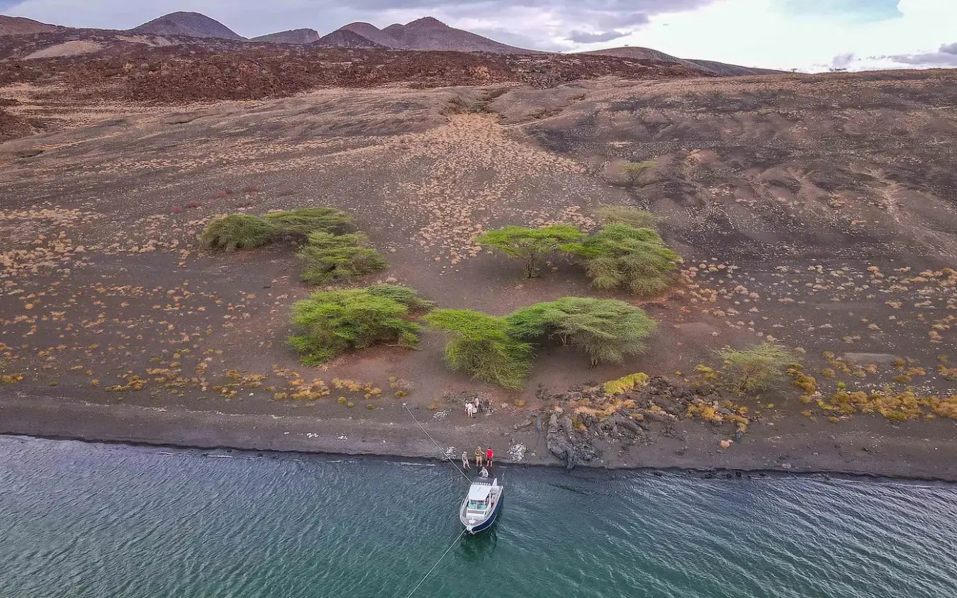 Small boat moored off volcanic black sand beach with trees, Koros Camp, Northern Kenya, Kenya