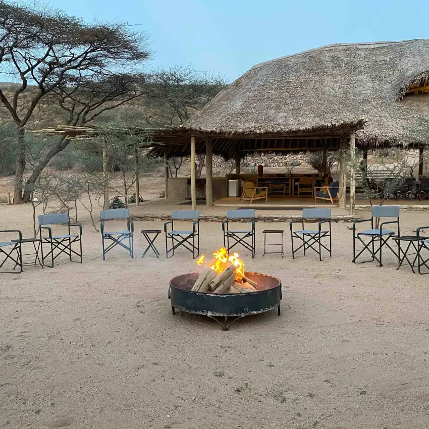 Central firepit with thatched-roof structures and camp chairs, Koros Camp, Northern Kenya, Kenya