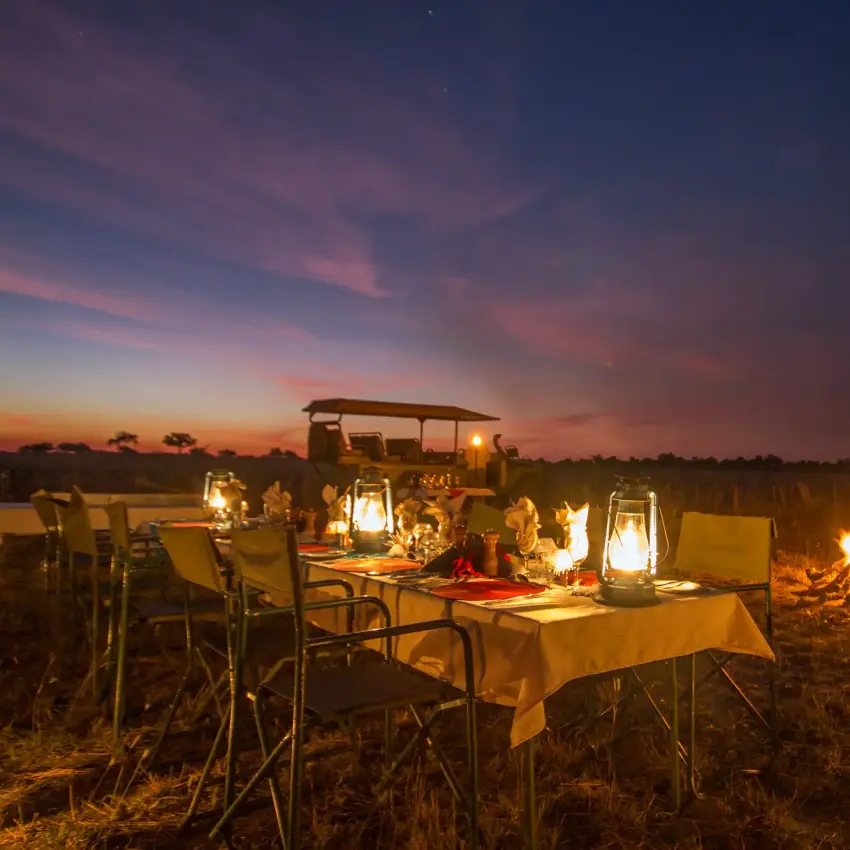 Dinner table set up in wilderness at dusk with lanterns and safari vehicle, Kwando Lagoon Camp, Linyanti Wildlife Reserve, Botswana