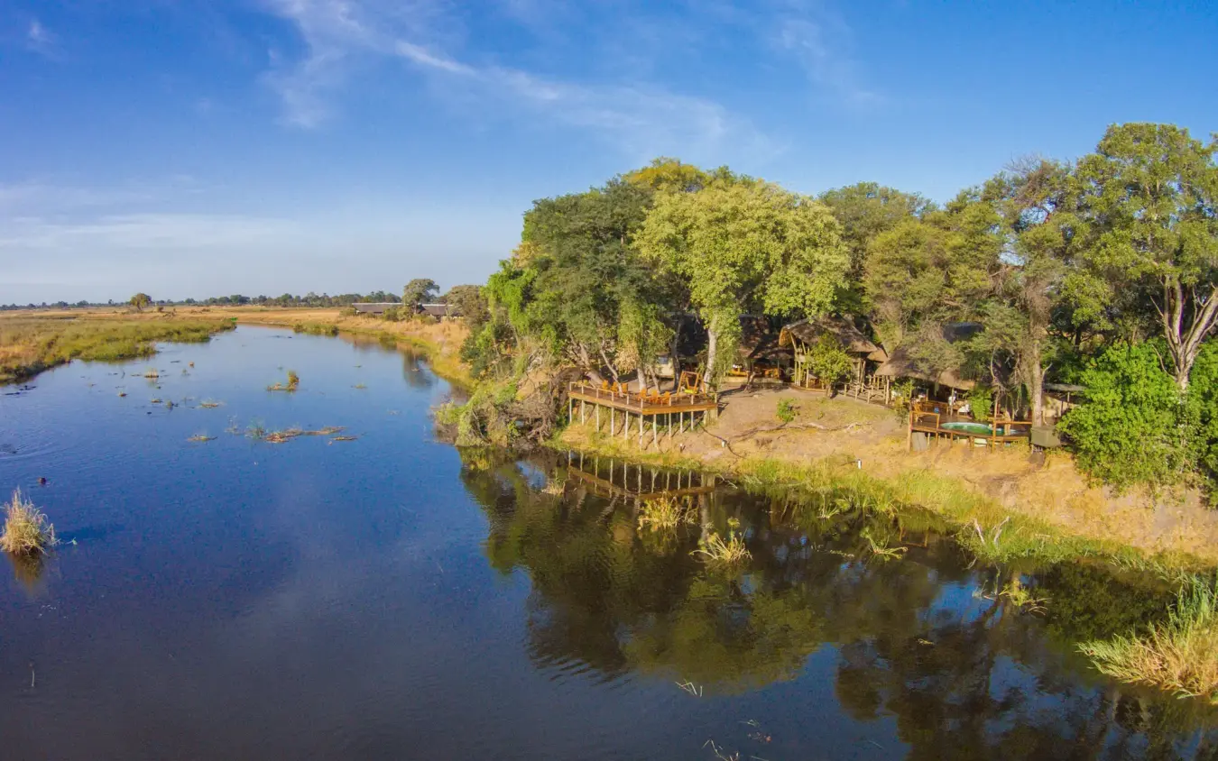Wooden boardwalk and safari lodge beside tranquil riverbank with trees, Kwando Lagoon Camp, Linyanti Wildlife Reserve, Botswana