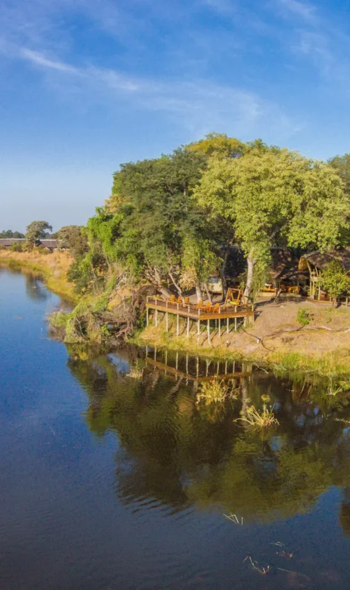 Wooden boardwalk and safari lodge beside tranquil riverbank with trees, Kwando Lagoon Camp, Linyanti Wildlife Reserve, Botswana