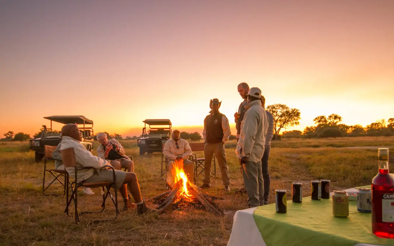 Guests gathered around campfire at sunset in open savanna landscape, Kwando Lebala Camp, Linyanti Wildlife Reserve, Botswana