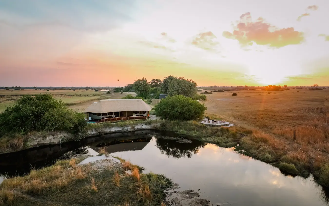 Thatched lodge beside waterhole at sunset with savanna grassland, Kwando Lebala Camp, Linyanti Wildlife Reserve, Botswana