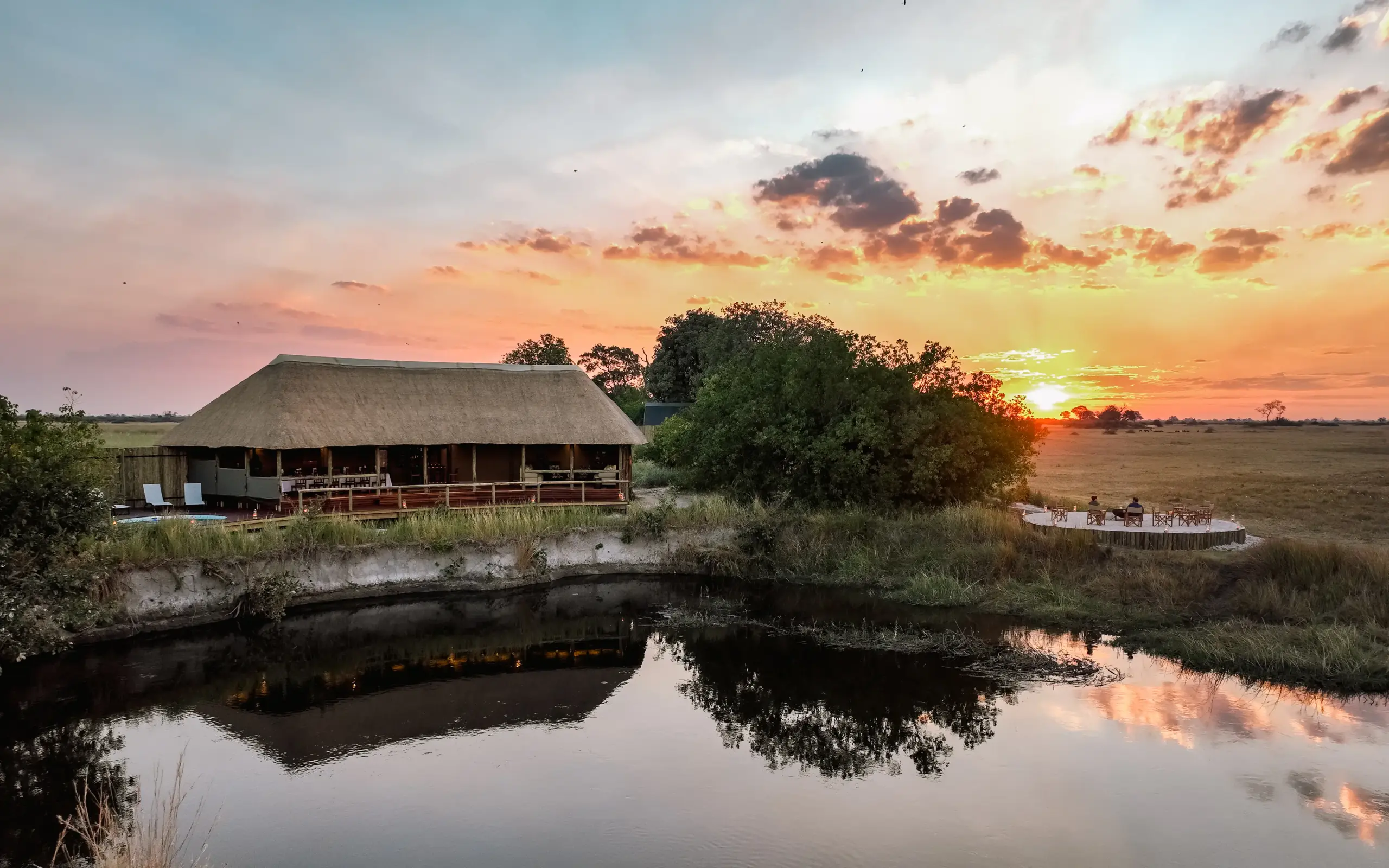 Thatched lodge beside water channel at sunset with boat moored nearby, Kwando Lebala Camp, Linyanti Wildlife Reserve, Botswana