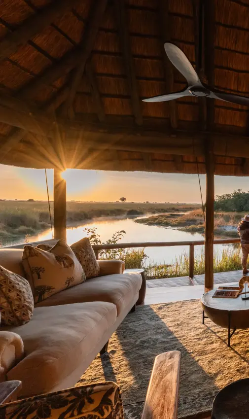 Open-sided lodge pavilion overlooking waterhole at sunset with guests gathered on deck, Kwando Lebala Camp, Linyanti Wildlife Reserve, Botswana
