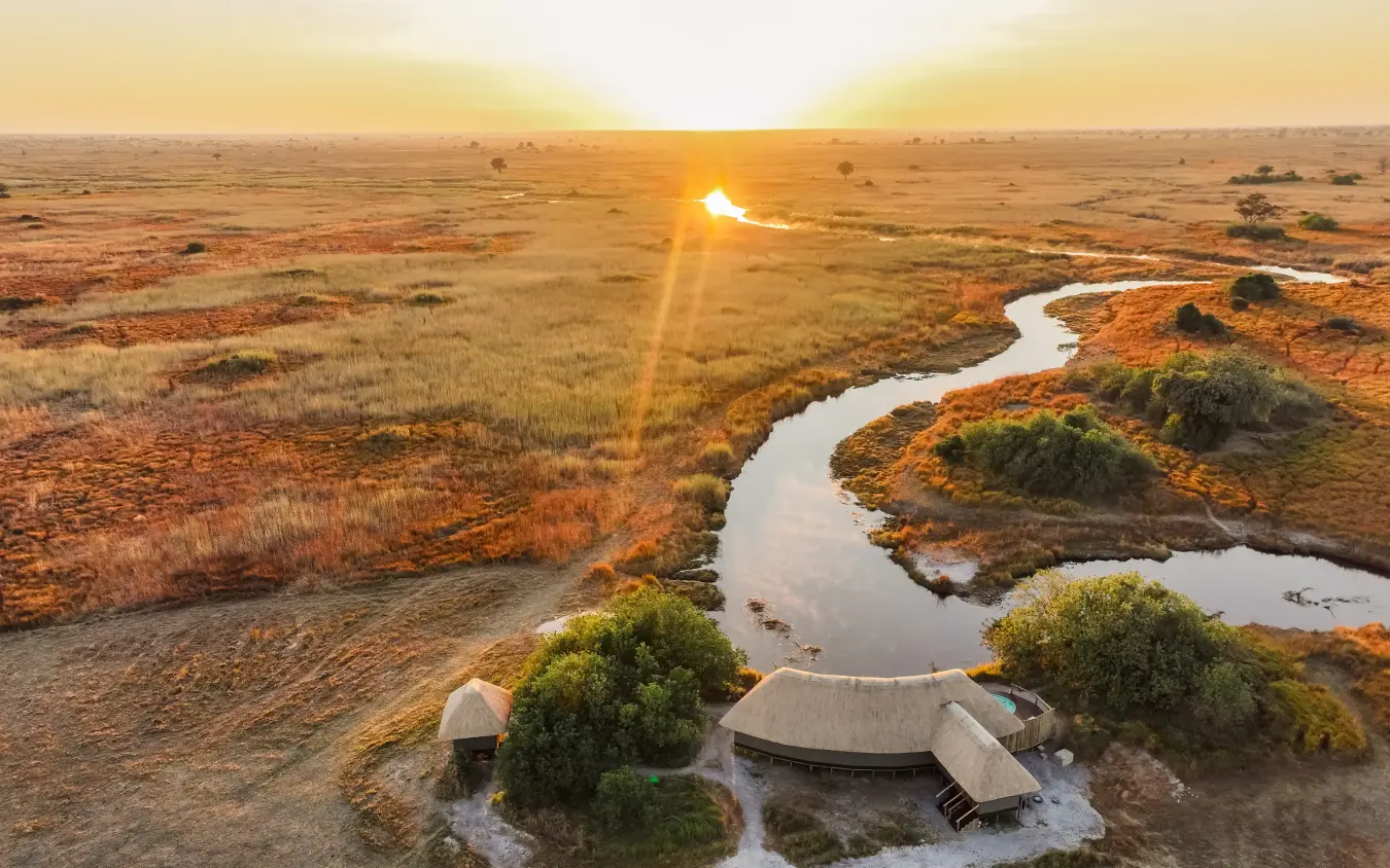 Thatched safari lodge beside winding river at sunset in open grassland, Kwando Lebala Camp, Linyanti Wildlife Reserve, Botswana