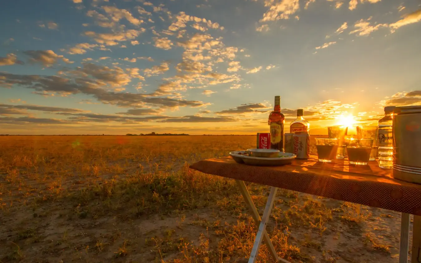 Outdoor dining setup with bottles and glasses at sunset in arid landscape, Kwando Nxai Pan Camp, Makgadikgadi Pans, Botswana