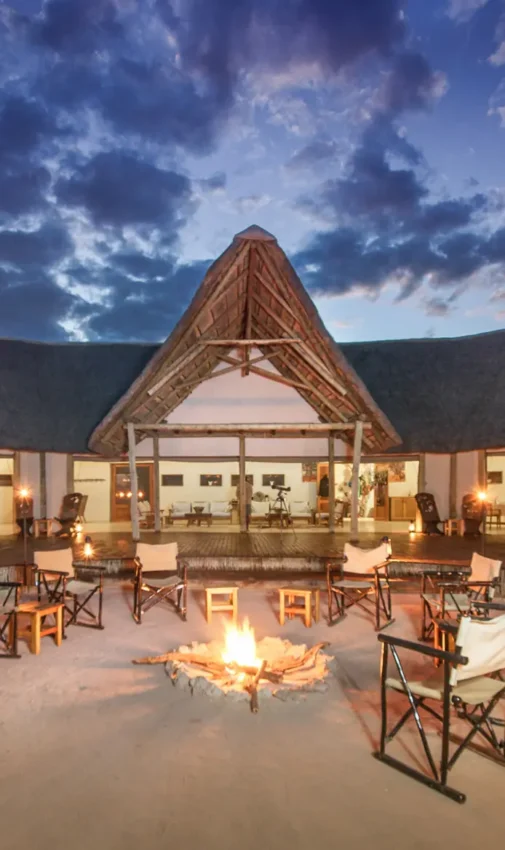 Luxury lodge with thatched roof and firepit seating on sandy ground at dusk, Kwando Nxai Pan Camp, Makgadikgadi Pans, Botswana