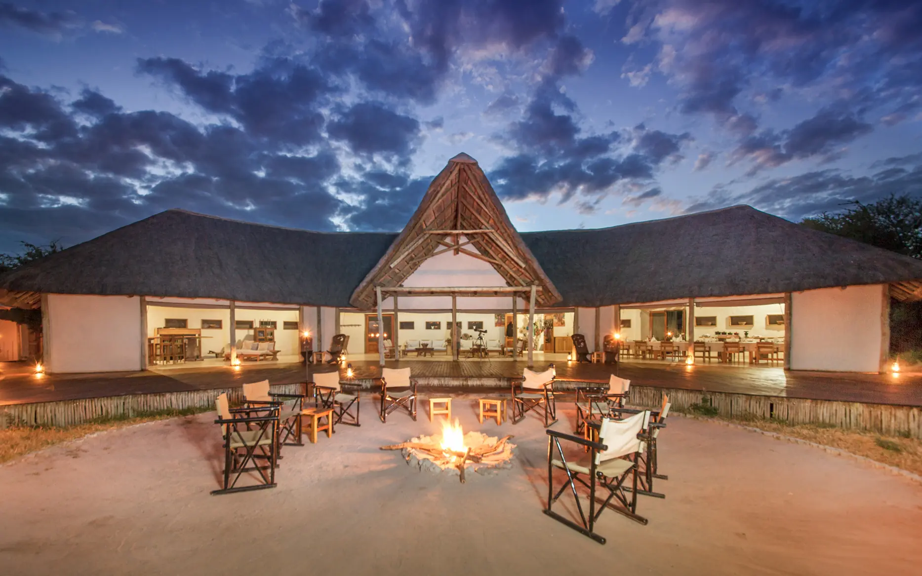 Luxury lodge with thatched roof and firepit seating on sandy ground at dusk, Kwando Nxai Pan Camp, Makgadikgadi Pans, Botswana