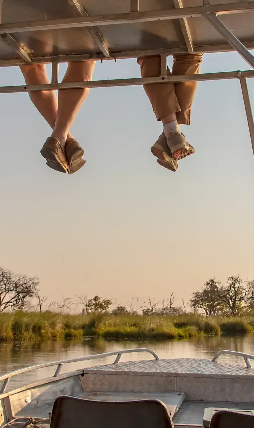 Boat with dangling feet on waterway at sunset, Kwando Splash Camp, The Okavango Delta, Botswana