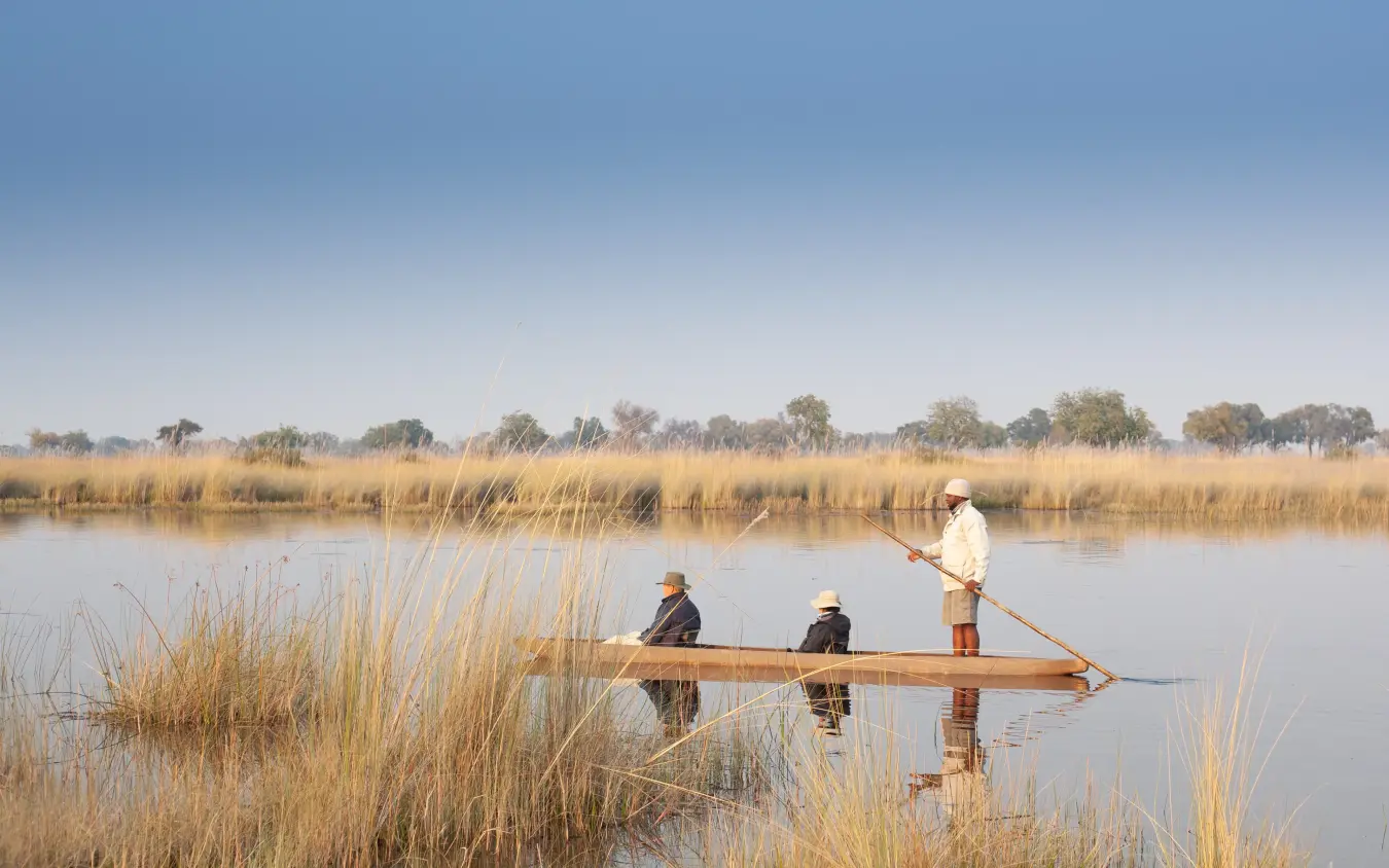 Local boatmen punting traditional wooden canoe through reed beds at waterhole, Kwando Splash Camp, The Okavango Delta, Botswana