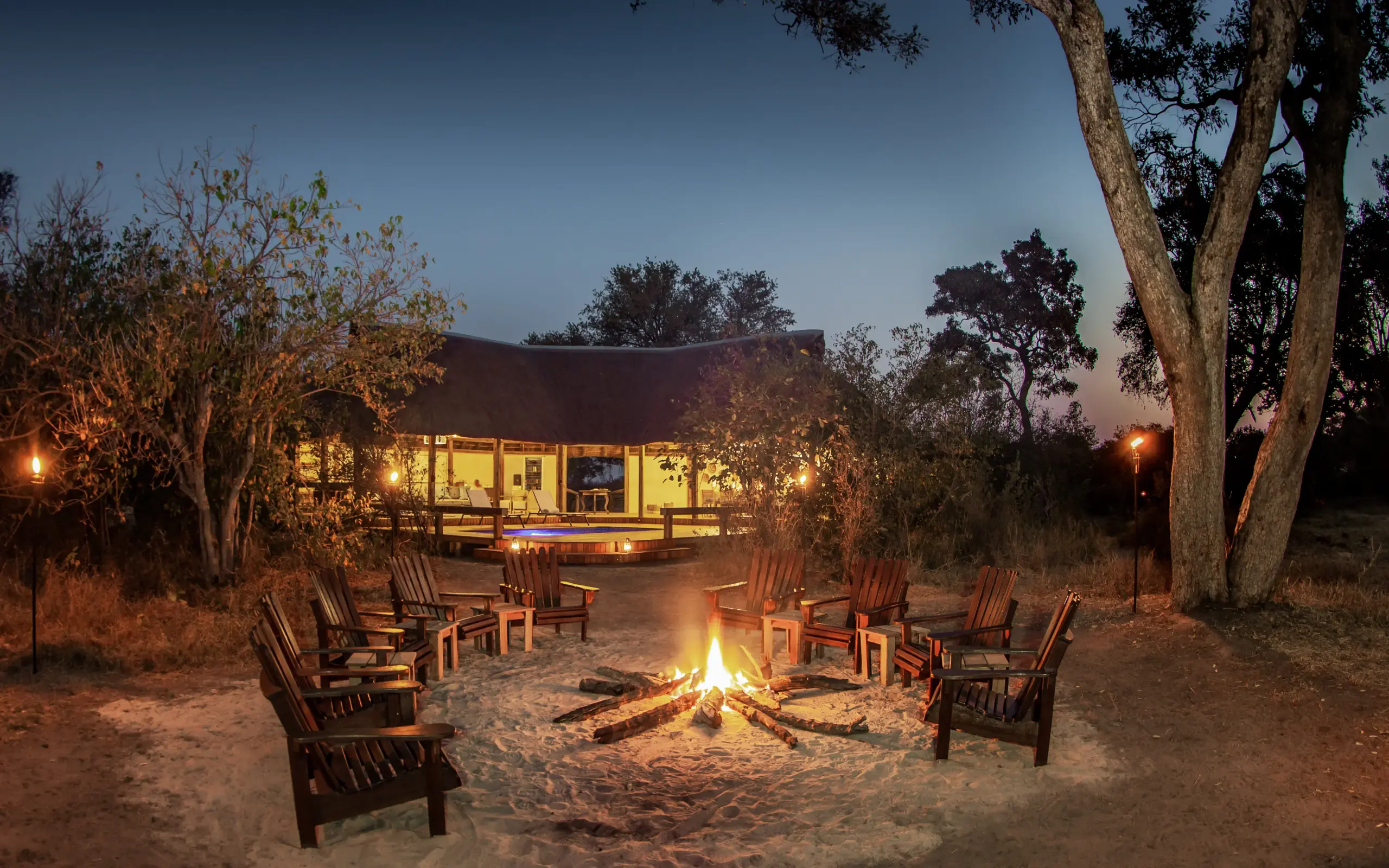 Campfire with Adirondack chairs around flames at dusk outside lit lodge, Kwando Splash Camp, The Okavango Delta, Botswana