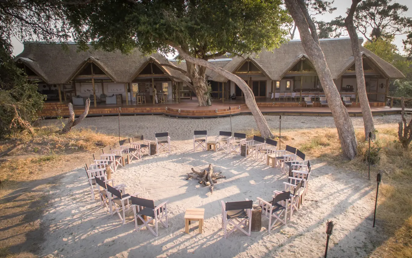 Circular seating arrangement around fire pit with safari lodge and thatched structures in background, Kwando Splash Camp, The Okavango Delta, Botswana