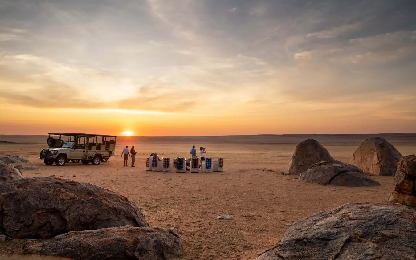 Safari vehicle with guests watching sunset over vast desert plain, Kwessi Dunes, NamibRand Private Reserve, Namibia