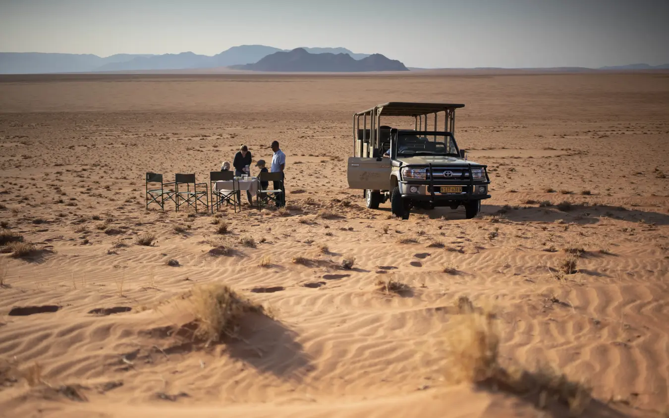 Safari vehicle with guests at desert camp beneath distant mountains, Kwessi Dunes, NamibRand Private Reserve, Namibia