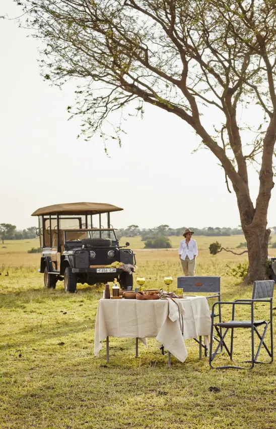 Outdoor dining setup with safari vehicle and acacia tree in grassland, Laba Grumeti Art Lodge, Serengeti National Park, Tanzania