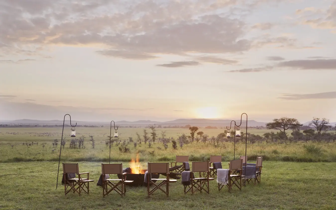Campfire with canvas chairs and lanterns in open grassland at sunset, Laba Migration Camp, Serengeti National Park, Tanzania