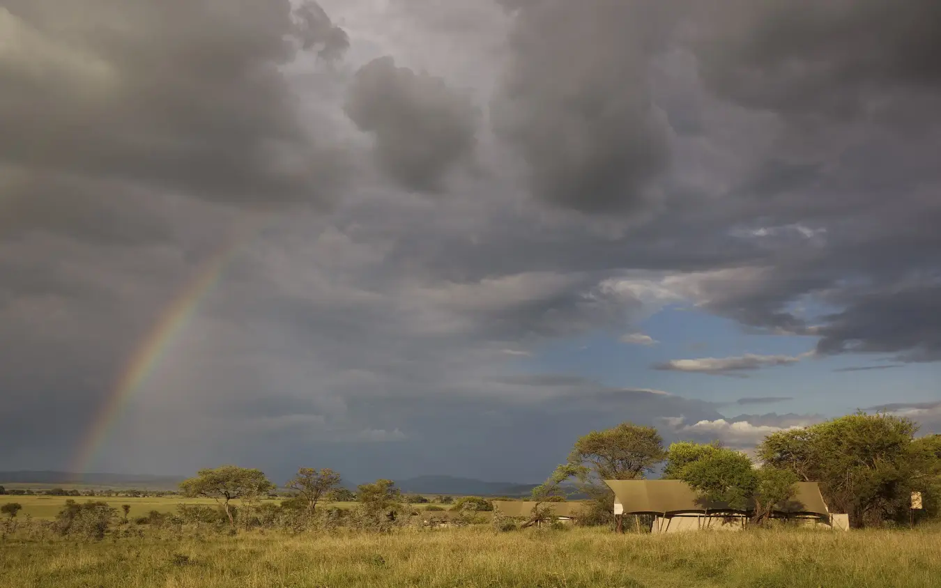Safari camp with canvas tent in open grassland under stormy sky with rainbow, Laba Migration Camp, Serengeti National Park, Tanzania