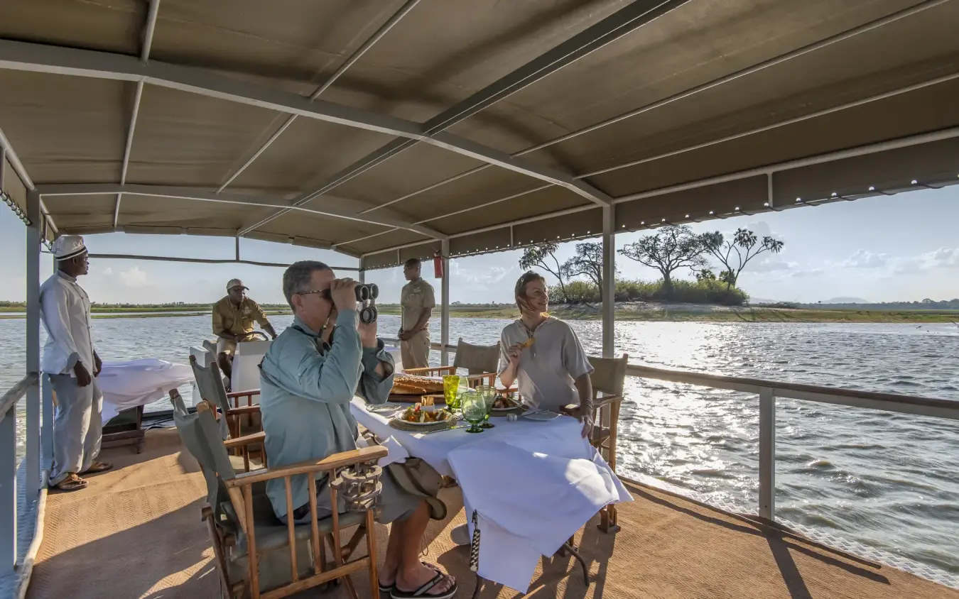 Guests dining on covered boat deck overlooking river wetland, Laba Siwandu, Nyerere National Park, Tanzania
