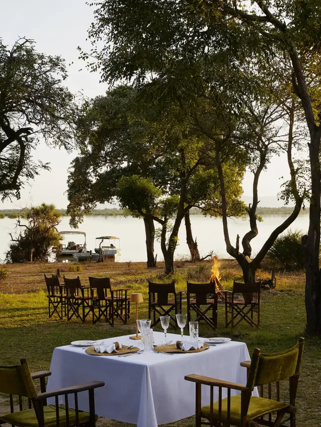 Outdoor dining setup with white tablecloth by river under acacia trees, Laba Siwandu, Nyerere National Park, Tanzania