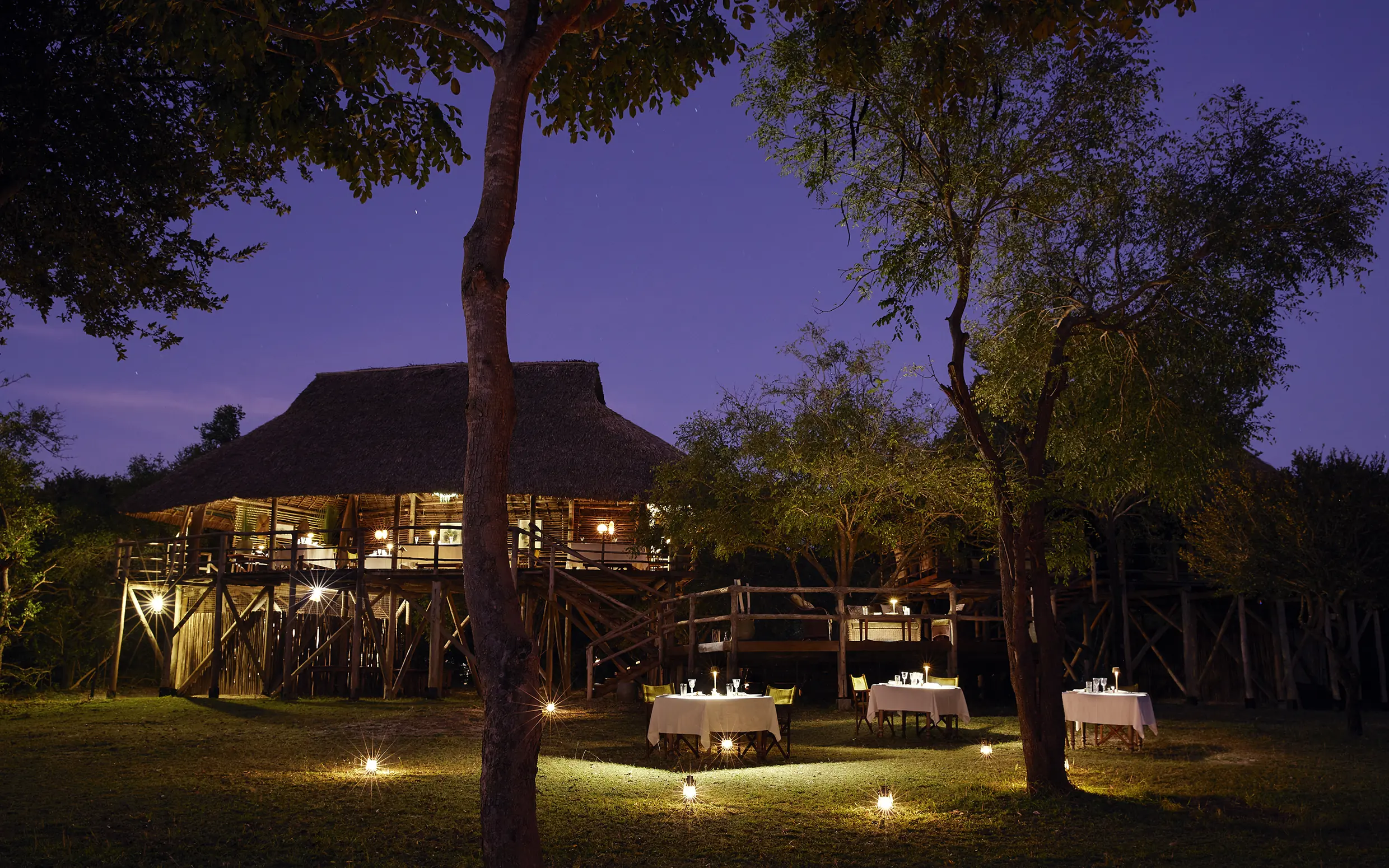 Illuminated lodge with thatched roof and wooden deck at dusk surrounded by trees, Laba Siwandu, Nyerere National Park, Tanzania