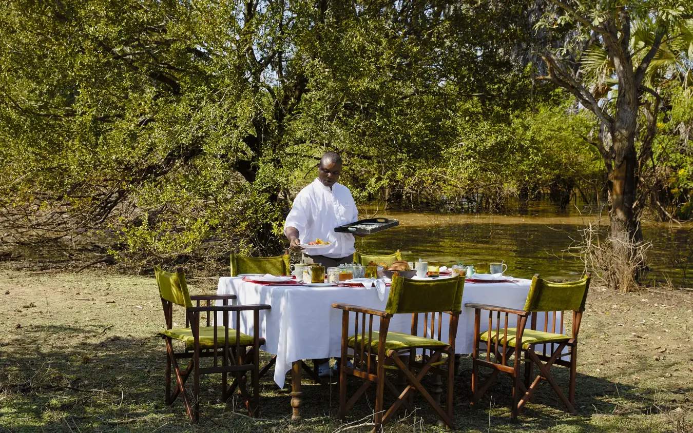 Outdoor dining setup by riverbank under leafy trees with server in white shirt, Laba Siwandu, Nyerere National Park, Tanzania
