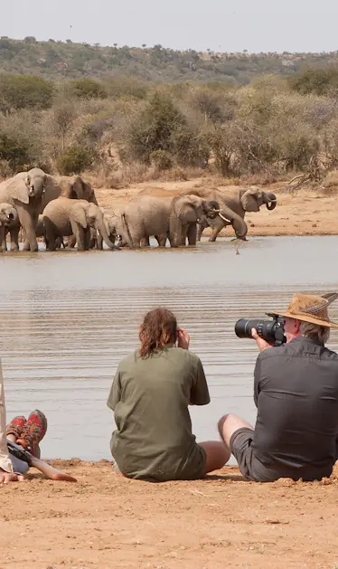 Guests photographing elephant herd at waterhole from shoreline, Laikipia Wilderness, Laikipia Highlands, Kenya
