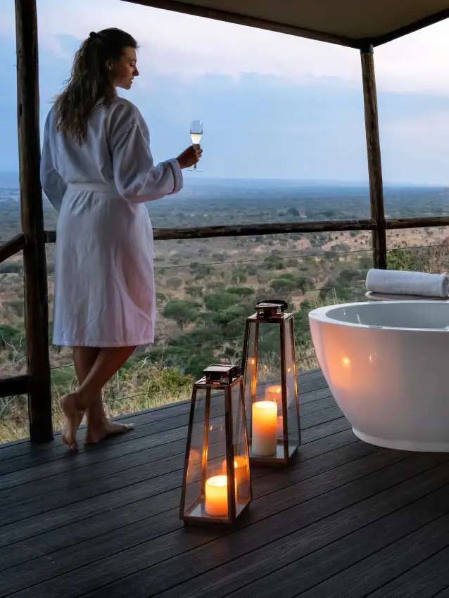 Woman in robe holding wine glass at elevated outdoor bathtub with savanna views, Mpingo Ridge Lodge, Tarangire National Park, Tanzania