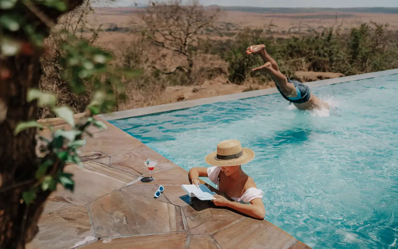 Guest relaxing poolside while another swims in infinity pool overlooking savanna landscape, Mpingo Ridge Lodge, Tarangire National Park, Tanzania