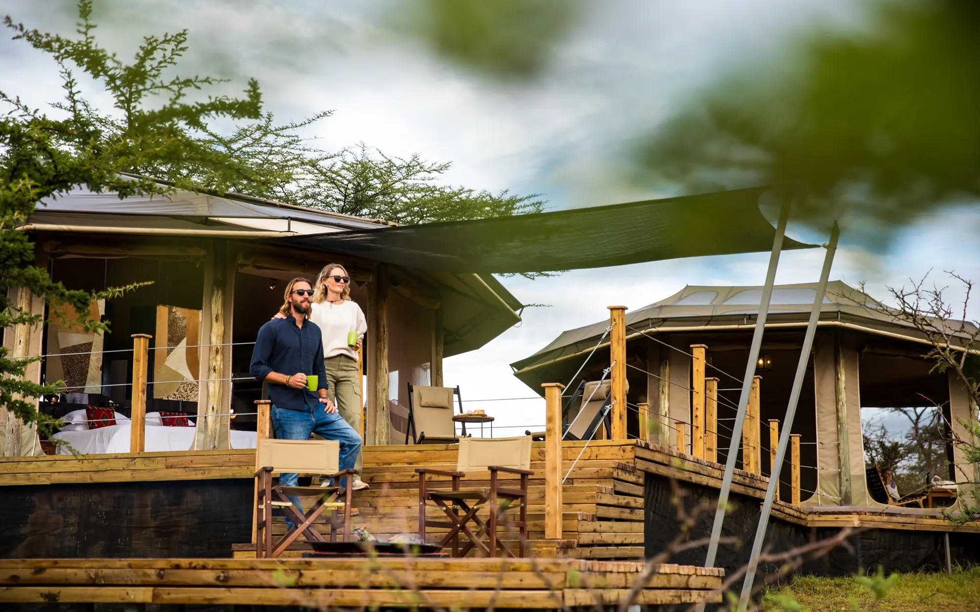 Guests on wooden safari lodge deck with shade sails and acacia trees, Leopard Hill, Maasai Mara, Kenya