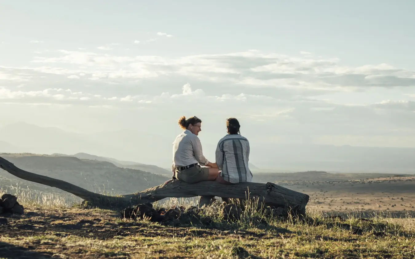 Two guests sitting on fallen log overlooking vast valley landscape, Lewa House, Laikipia Highlands, Kenya