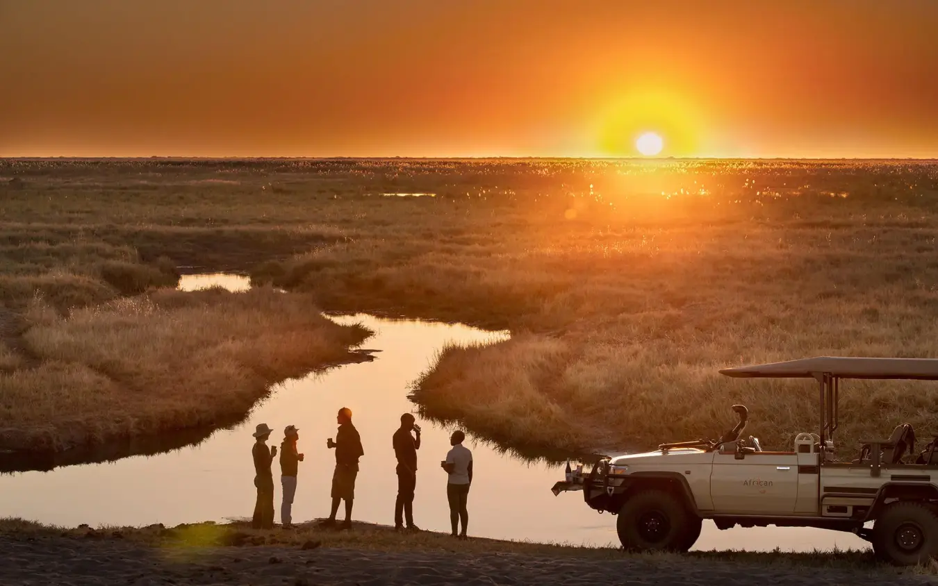 Guests standing by safari vehicle at sunset overlooking wetland, Linyanti Bush Camp, Linyanti Wildlife Reserve, Botswana