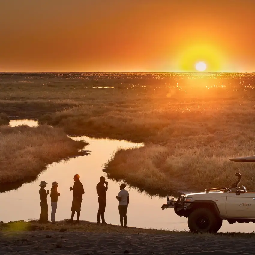 Guests standing by safari vehicle at sunset overlooking wetland, Linyanti Bush Camp, Linyanti Wildlife Reserve, Botswana