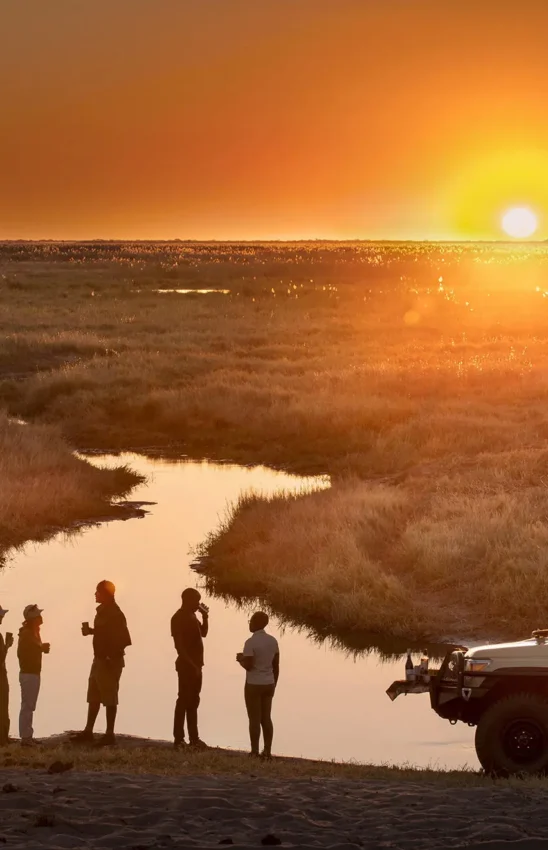 Guests standing beside safari vehicle at sunset overlooking wetland, Linyanti Wildlife Reserve, Botswana