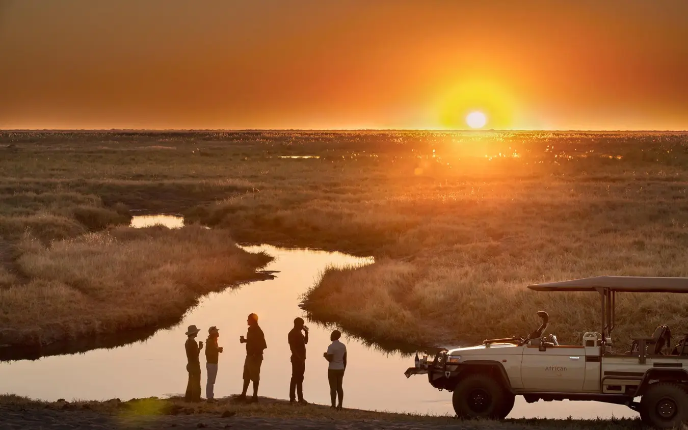 Guests standing beside safari vehicle at sunset overlooking wetland, Linyanti Ebony, Linyanti Wildlife Reserve, Botswana