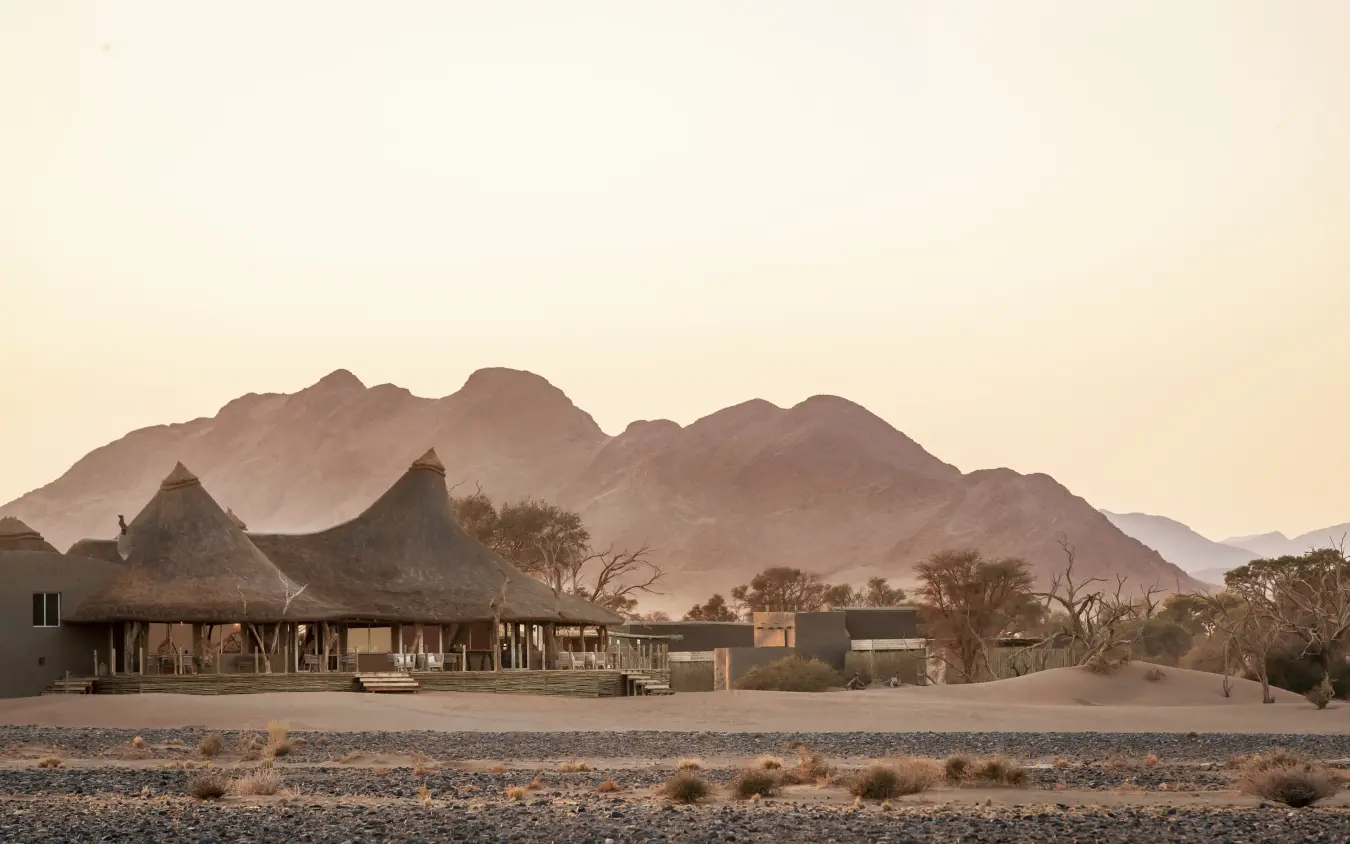 Desert lodge with thatched roofs backed by dramatic mountain peaks at dusk, Little Kulala, Sossusvlei, Namibia