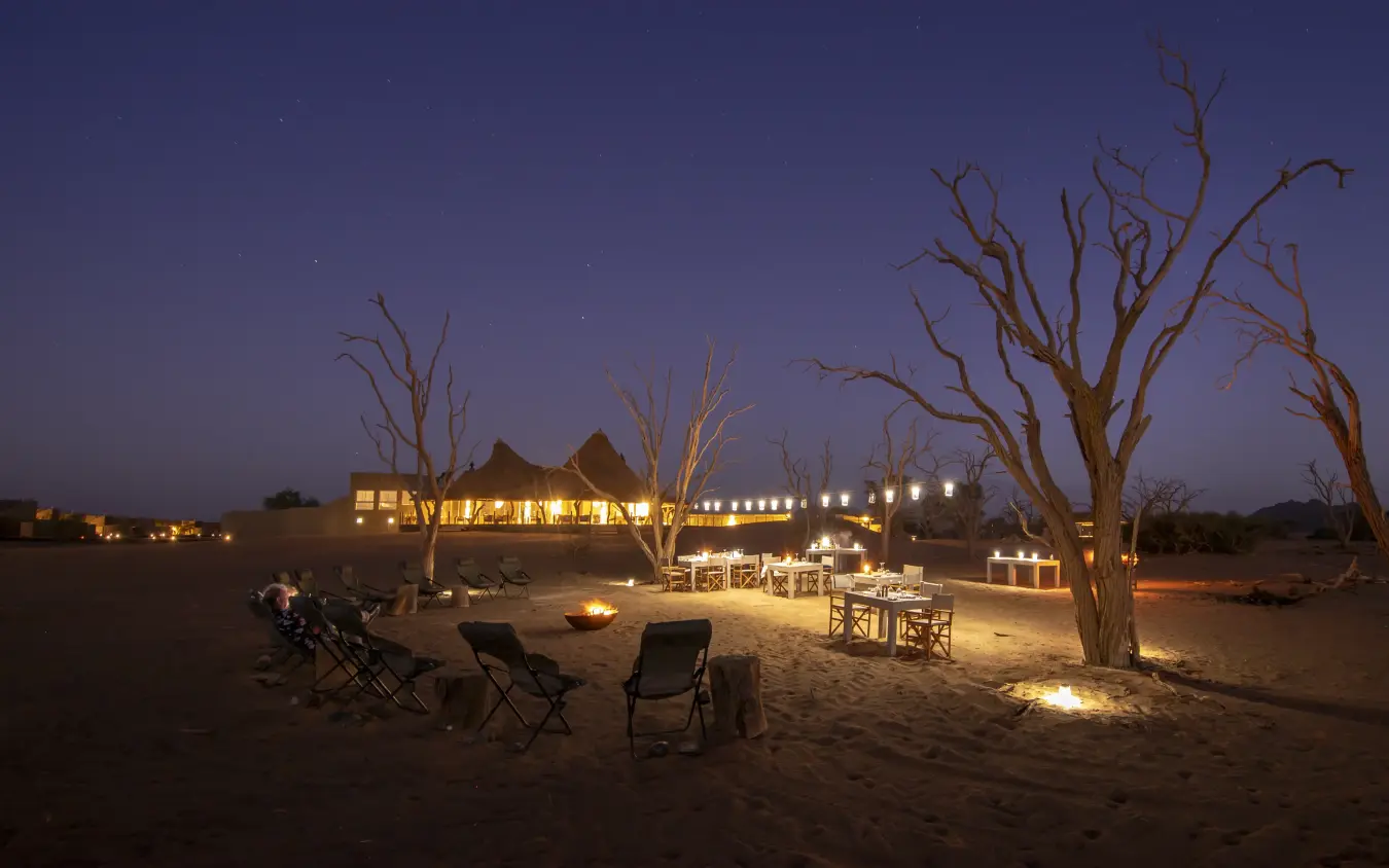 Luxury lodge illuminated at night with bare trees and campfire seating in desert, Little Kulala, Sossusvlei, Namibia