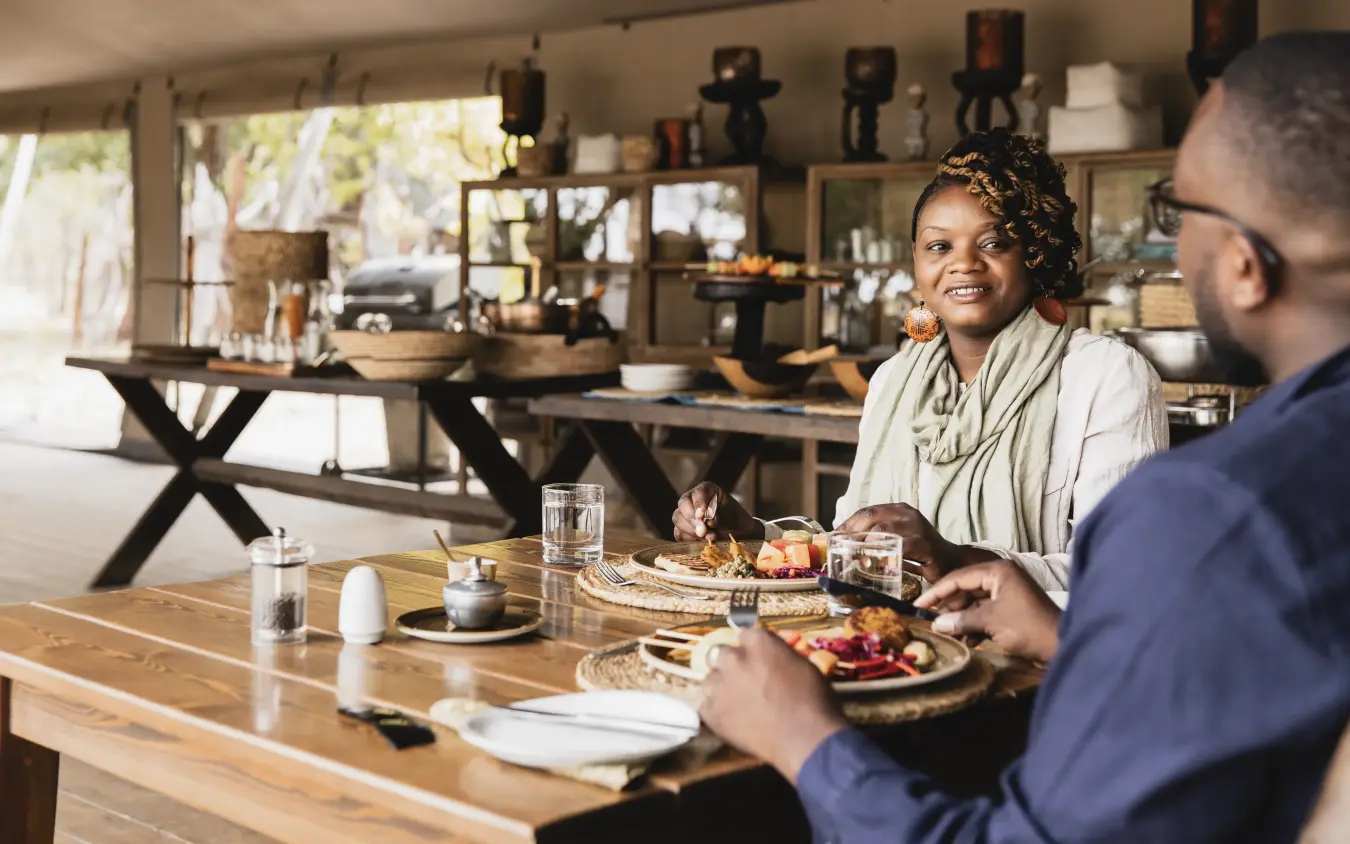 Couple dining at wooden table in open-sided safari lodge, Little Makalolo, Hwange National Park, Zimbabwe
