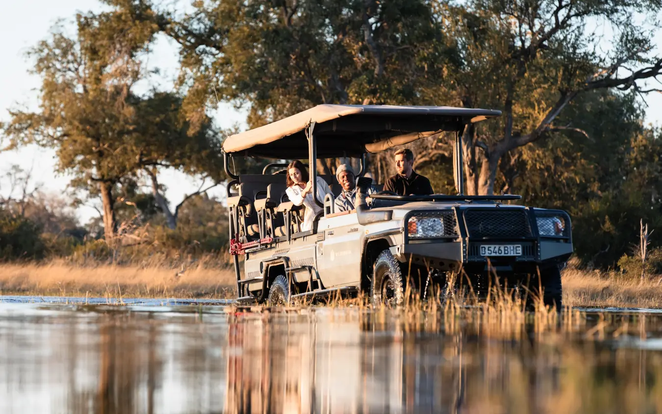 Safari vehicle with guests crossing shallow water at golden hour, Little Vumbura, The Okavango Delta, Botswana