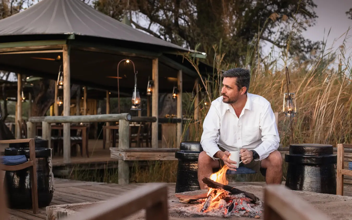 Man sitting by campfire at luxury safari lodge with tented accommodation, Little Vumbura, The Okavango Delta, Botswana
